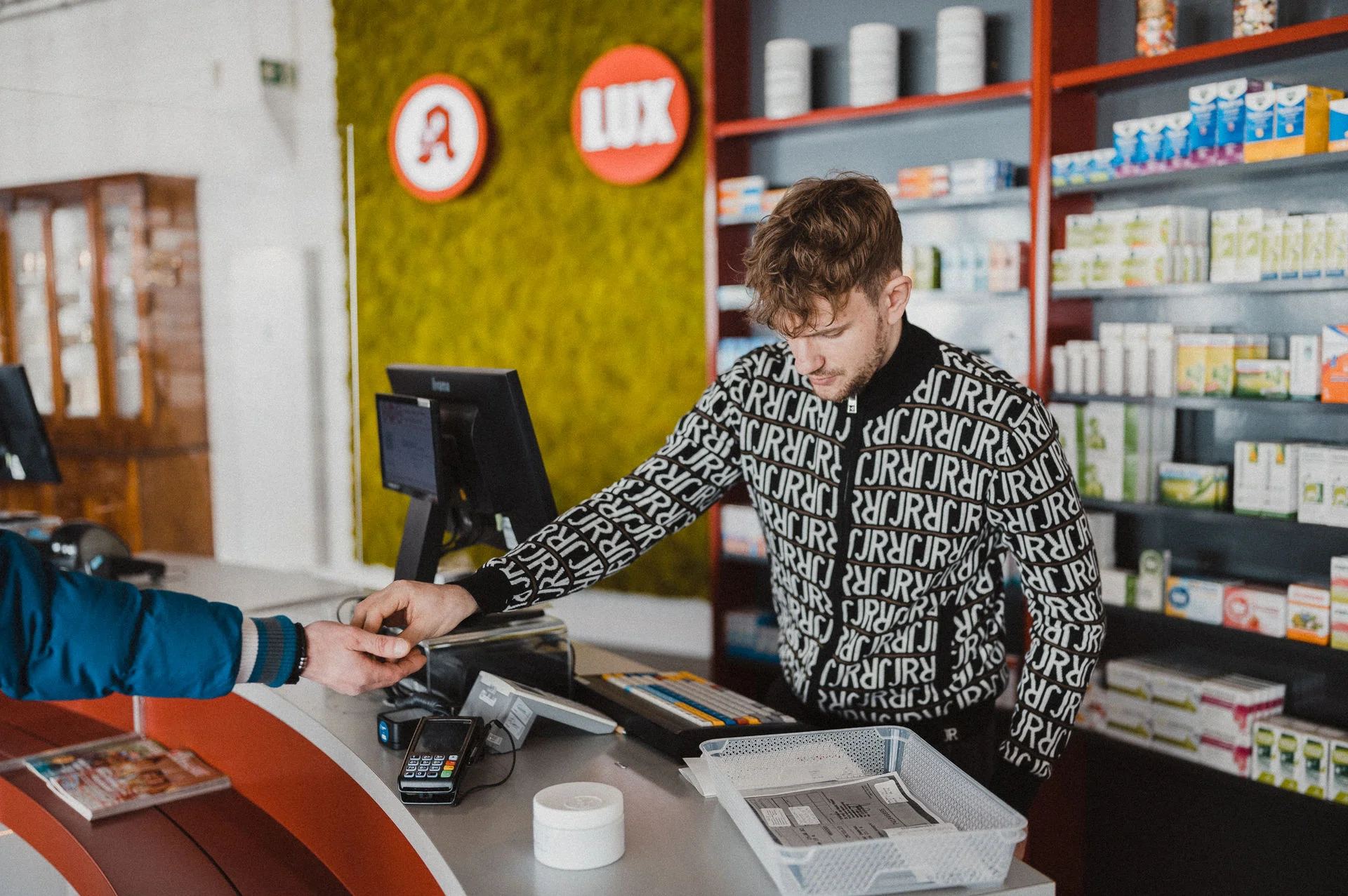 Szene im Verkaufsbereich der Apotheke: Am Handverkaufstisch erfolgt die persönliche Beratung und Übergabe von Arzneimitteln. Im Hintergrund sind die Sichtwahlregale mit verschiedenen Gesundheits- und Apothekenprodukten zu sehen. Die Aufnahme vermittelt den direkten und serviceorientierten Kontakt zwischen Apotheke und Kundschaft.