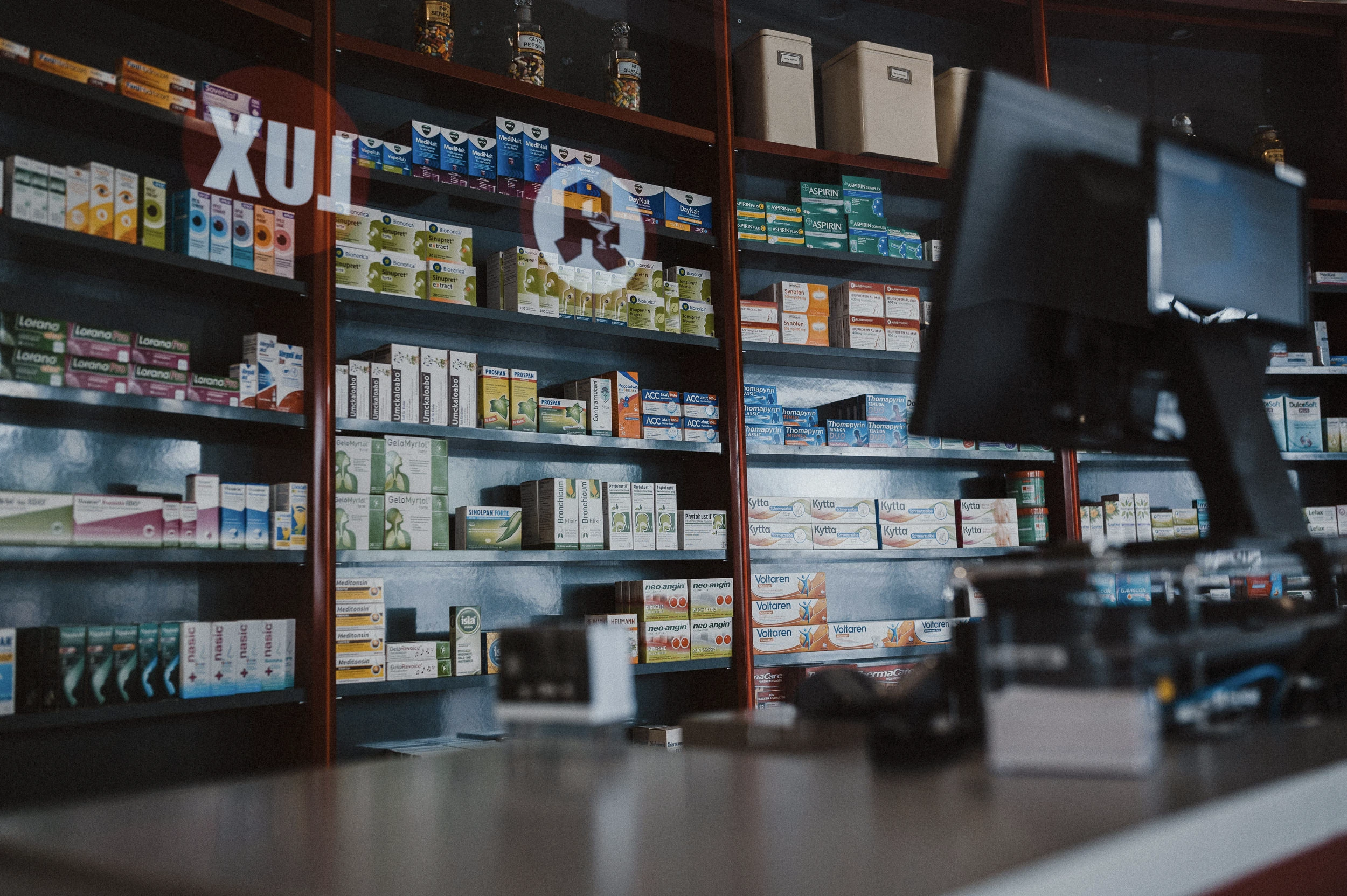 Innenansicht des Verkaufsraums der Apotheke. Im Hintergrund befinden sich gut sortierte Sichtwahlregale mit Arzneimitteln und Gesundheitsprodukten. Im Vordergrund ist der Handverkaufstisch mit Kassensystemen zu sehen, an dem die persönliche Beratung und Abgabe von Medikamenten erfolgt.